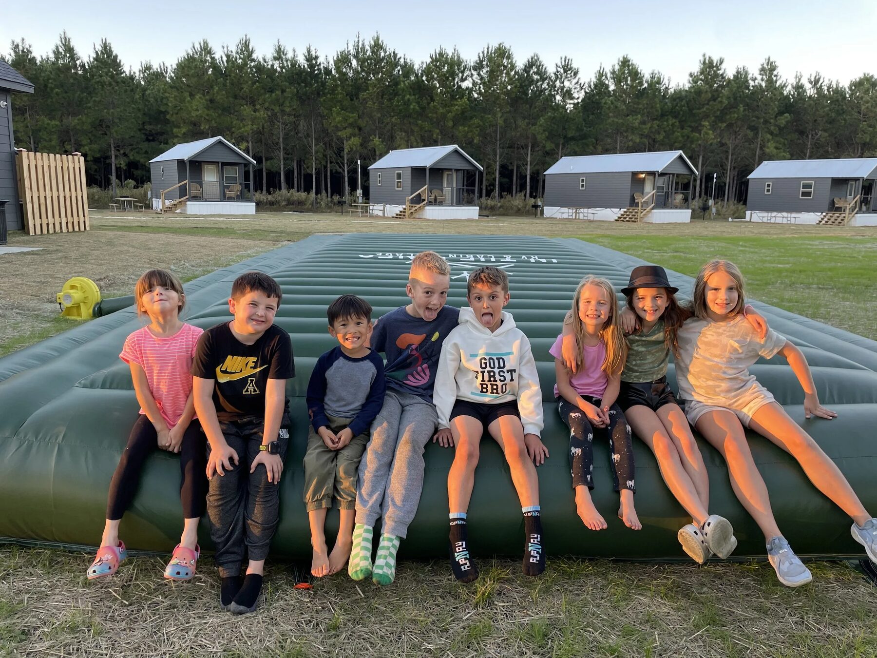 Nine children sitting in a row along the jump pad at In The Pines, cabins and pines behind them